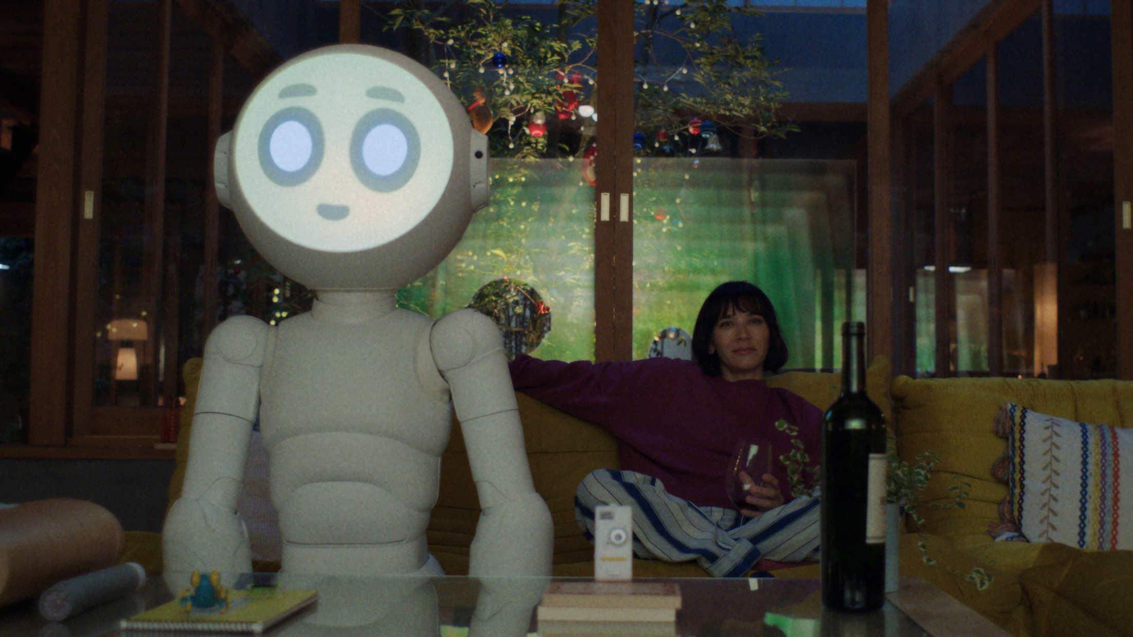 A white robot with a large face watches TV next to a woman sitting on a couch in a modern looking home.