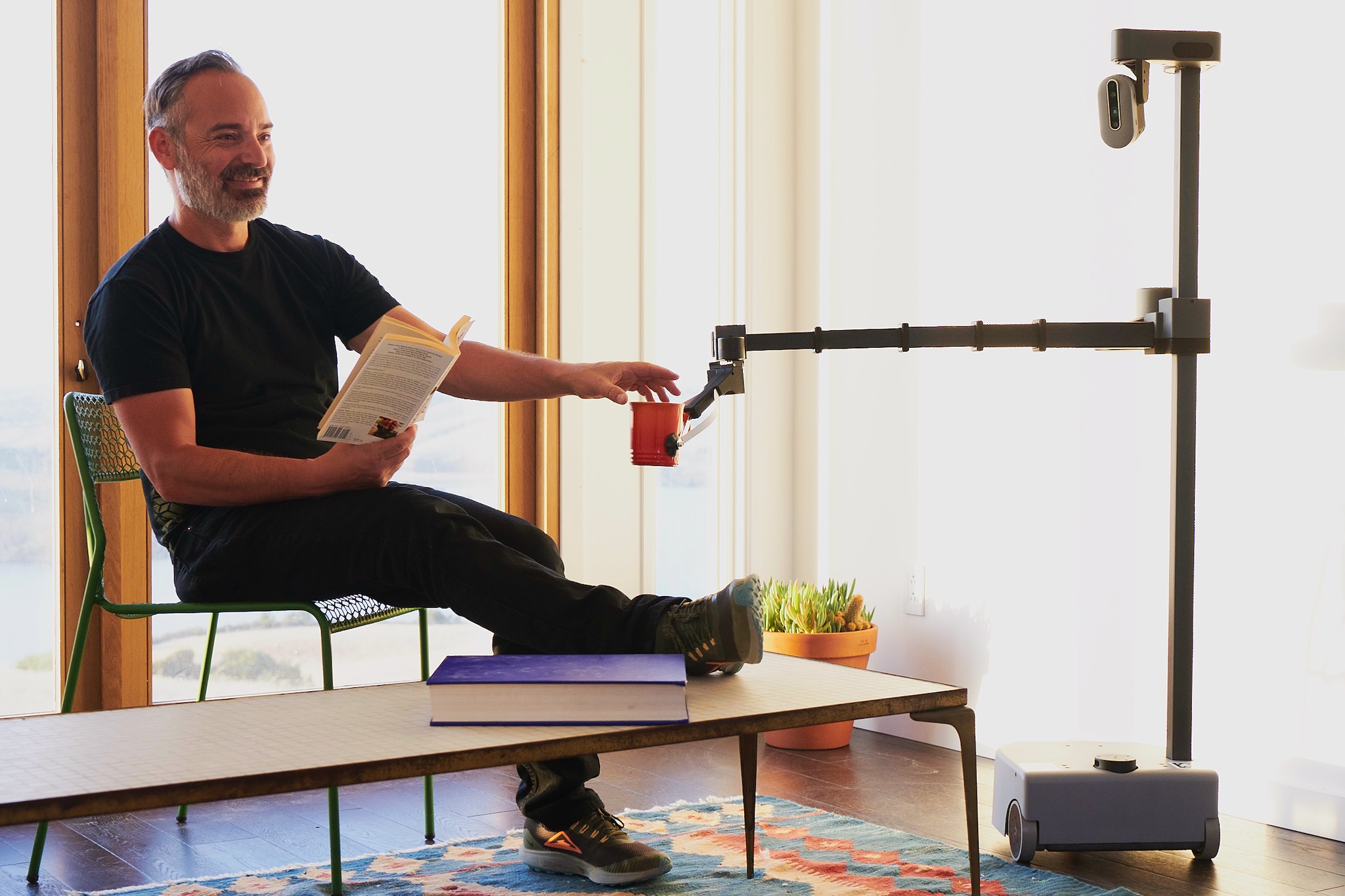 A robot consisting of a mobile base that carries a pole with a camera and an arm delivers a cup to a smiling man sitting on a chair while reading a book in a living room.