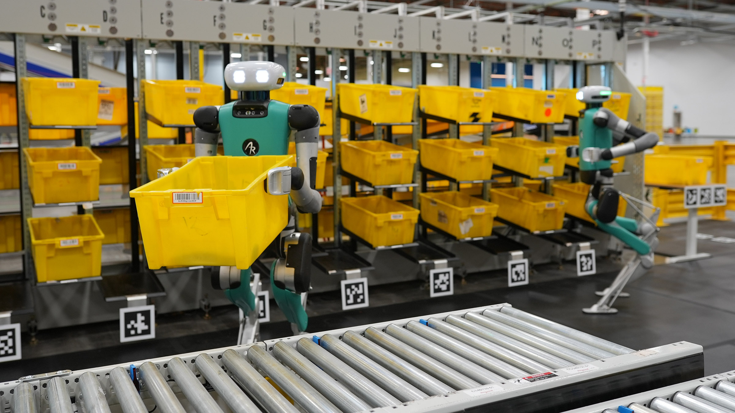 Two Digit humanoid robots with green and gray bodies carry yellow plastic bins in a warehouse.