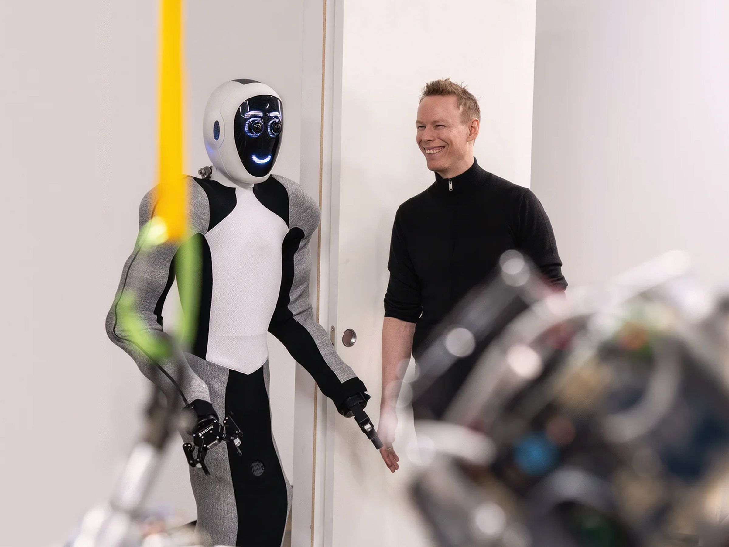 Humanoid robot Eve, with torso and arms covered by fabric, wheels for feet, and a smiling screen face, stands next to a smiling man with spiky hair wearing a black shirt.