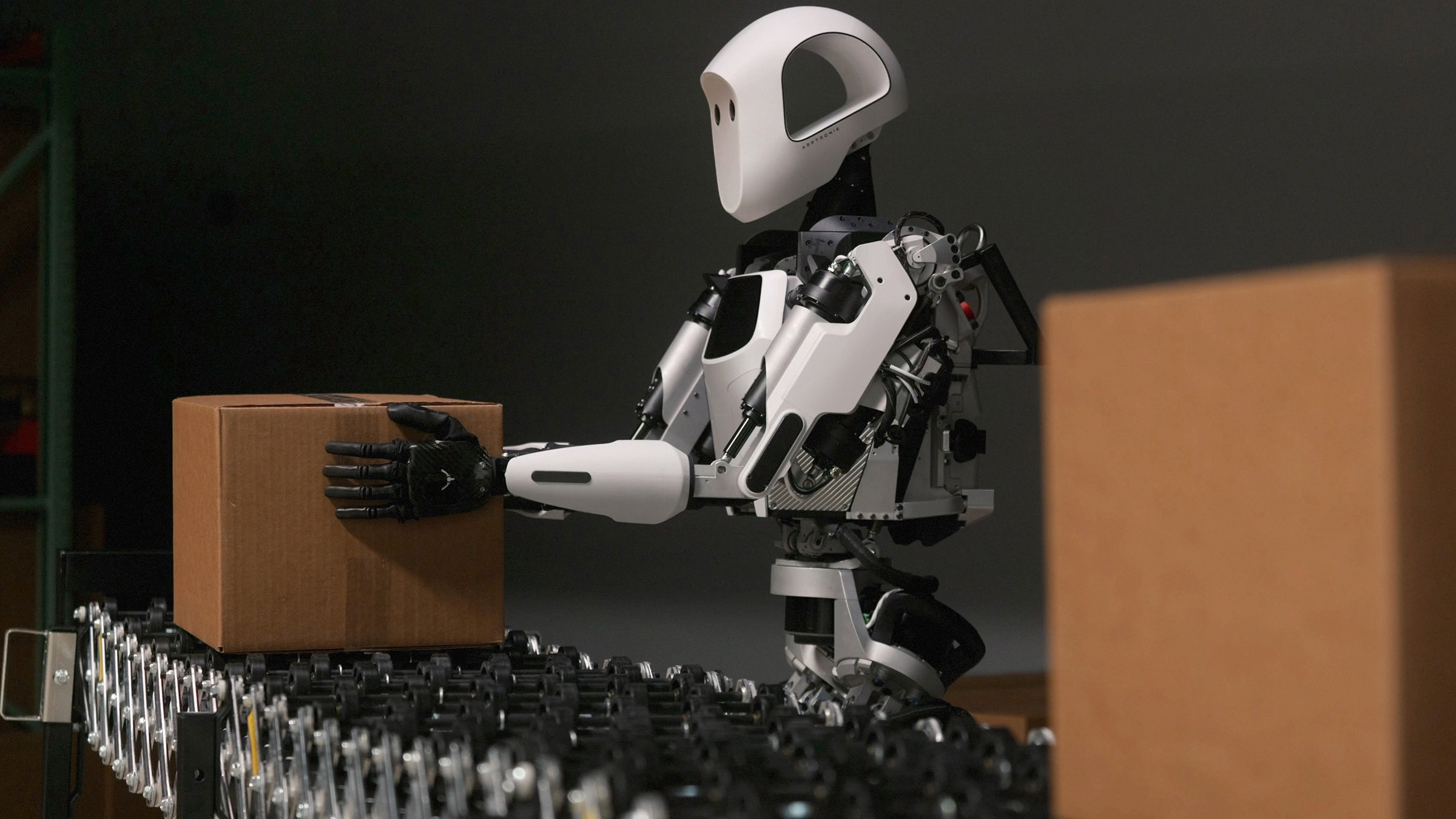 A humanoid robot the size of a person picks up a cardboard box from a conveyor belt.