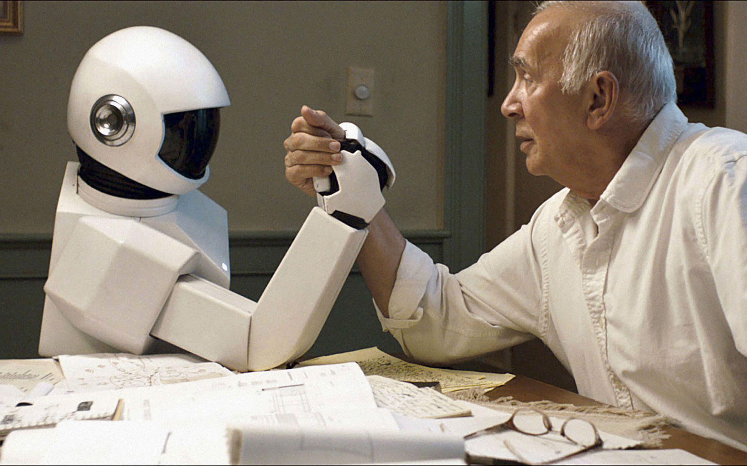 A white humanoid robot arm wrestles a man with white hair on a kitchen table.