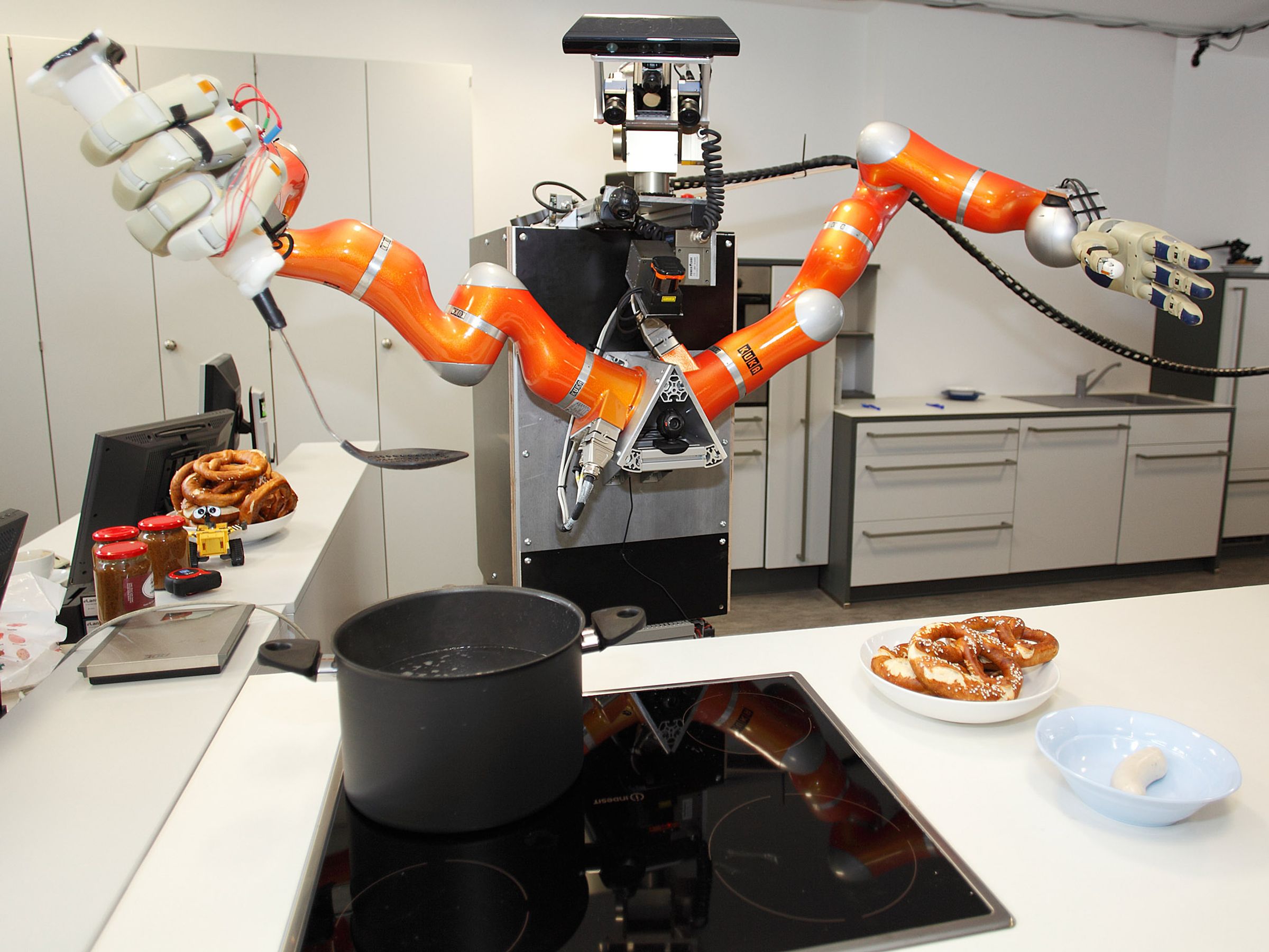 A tall humanoid robot with orange arms and large robotic hands removes sausages from boiling water in a kitchen.