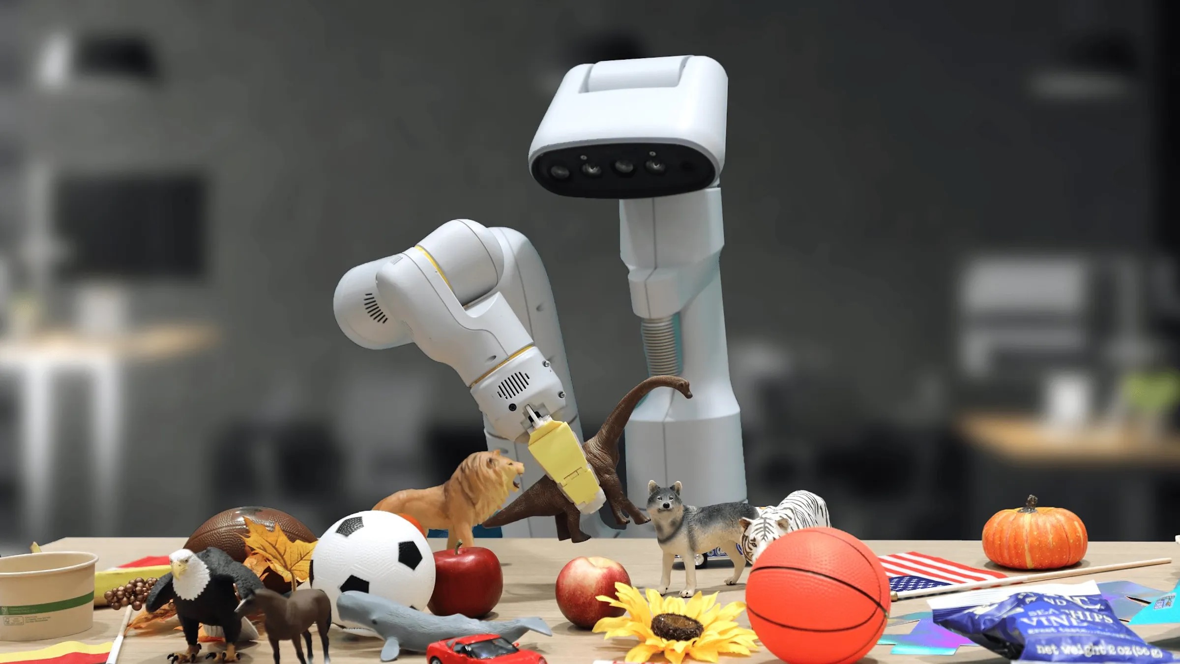 A white robot with a long neck, camera head, and one arm, holds a little dinosaur toy in its gripper.