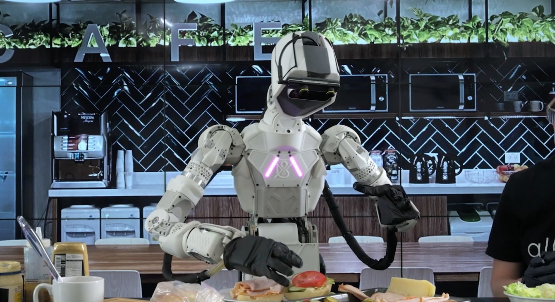 A teleoperated humanoid robot looks down while using its hands to assemble a turkey sandwich in a kitchen laboratory.