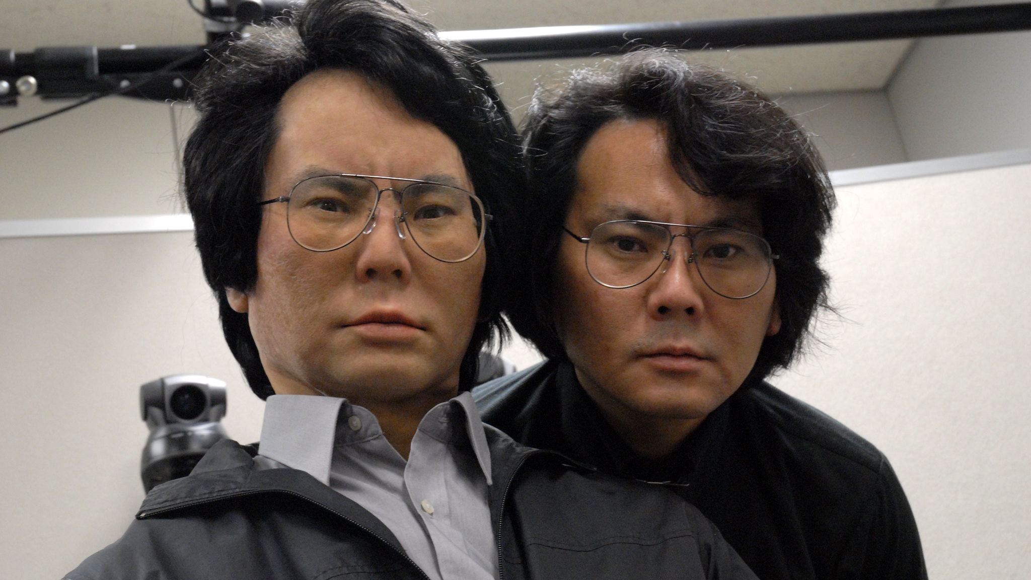 Japanese roboticist Hiroshi Ishiguro, with dark hair and glasses, stands next to his android twin Geminoid HI-1, which has the same hair and glasses.